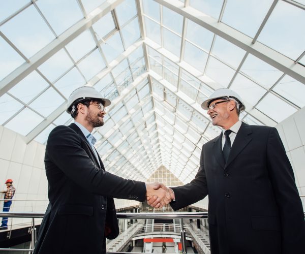 Two cheerful businessman handshaking in helmet at new building.